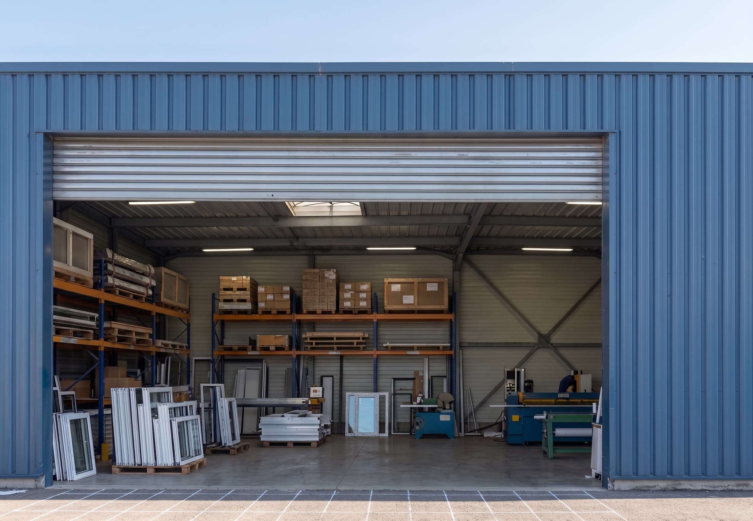 Intérieur de l'atelier Atelier Fermetures vu depuis la porte : stock de profilés, machines et commandes palettisées prêtes au départ
